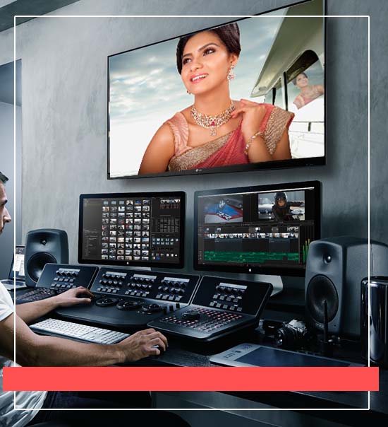 A person is working in a video editing studio, sitting at a control panel with multiple screens in front of them. The main screen displays a woman in traditional Indian attire, wearing a saree and jewelry. The other screens show various editing software interfaces, indicating that the person is engaged in video post-production work. The room has a professional and high-tech atmosphere.