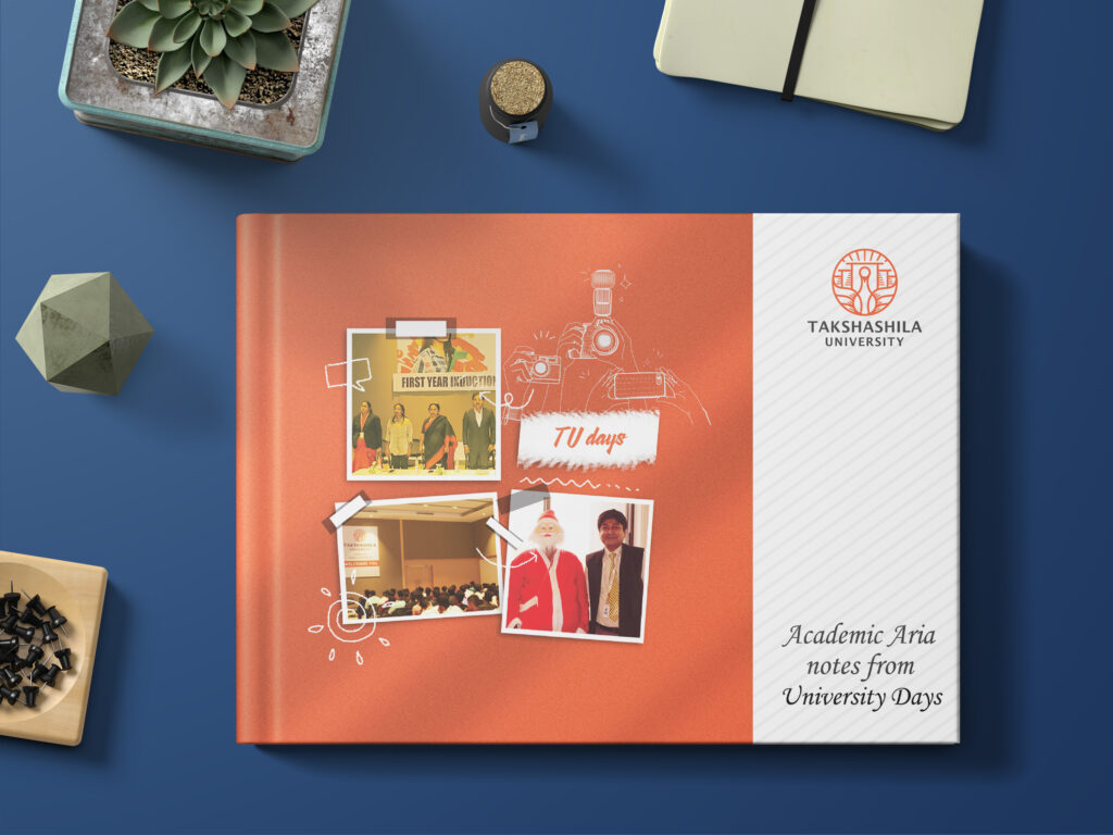 The image shows a flat lay of a photo album titled "Academic Aria notes from University Days" with a logo of "Takshashila University" on the front cover. The album has a color scheme of orange and white, with small photo printouts showing university-related events like "First Year Induction." Decorative doodles, such as a camera and polaroid frames, are drawn around the photos. The album is placed on a dark blue desk surface, surrounded by objects like a succulent plant, a notepad, binder clips, and a geometric paperweight.
