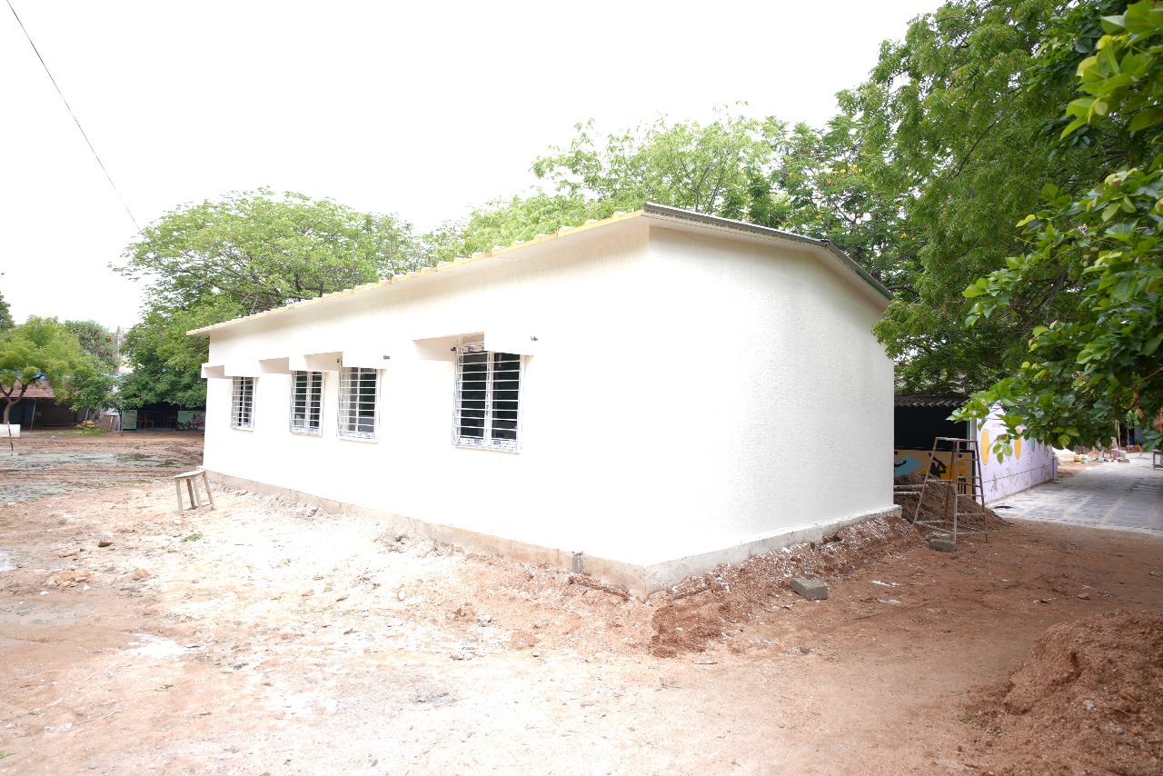 A single-story prefabricated house, painted white with several windows and metal bars, stands on bare ground surrounded by trees under a bright sky.
